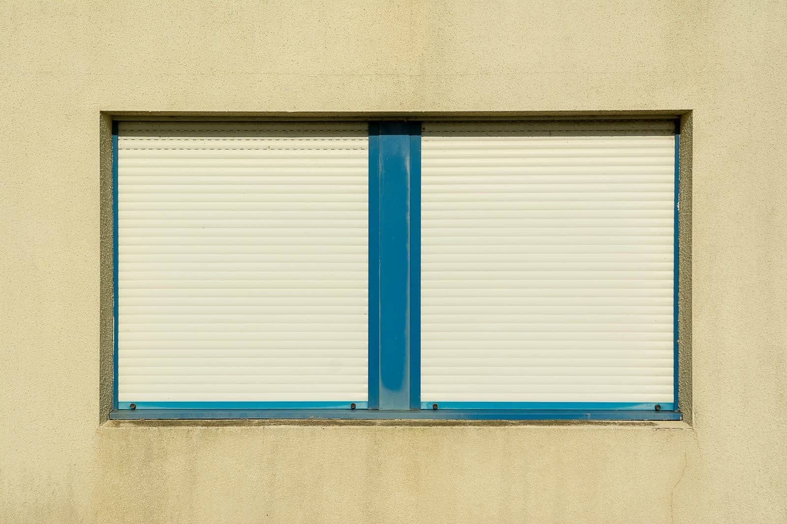 Dos ventanas en una pared blanca, una con la persiana a media altura y otra subida